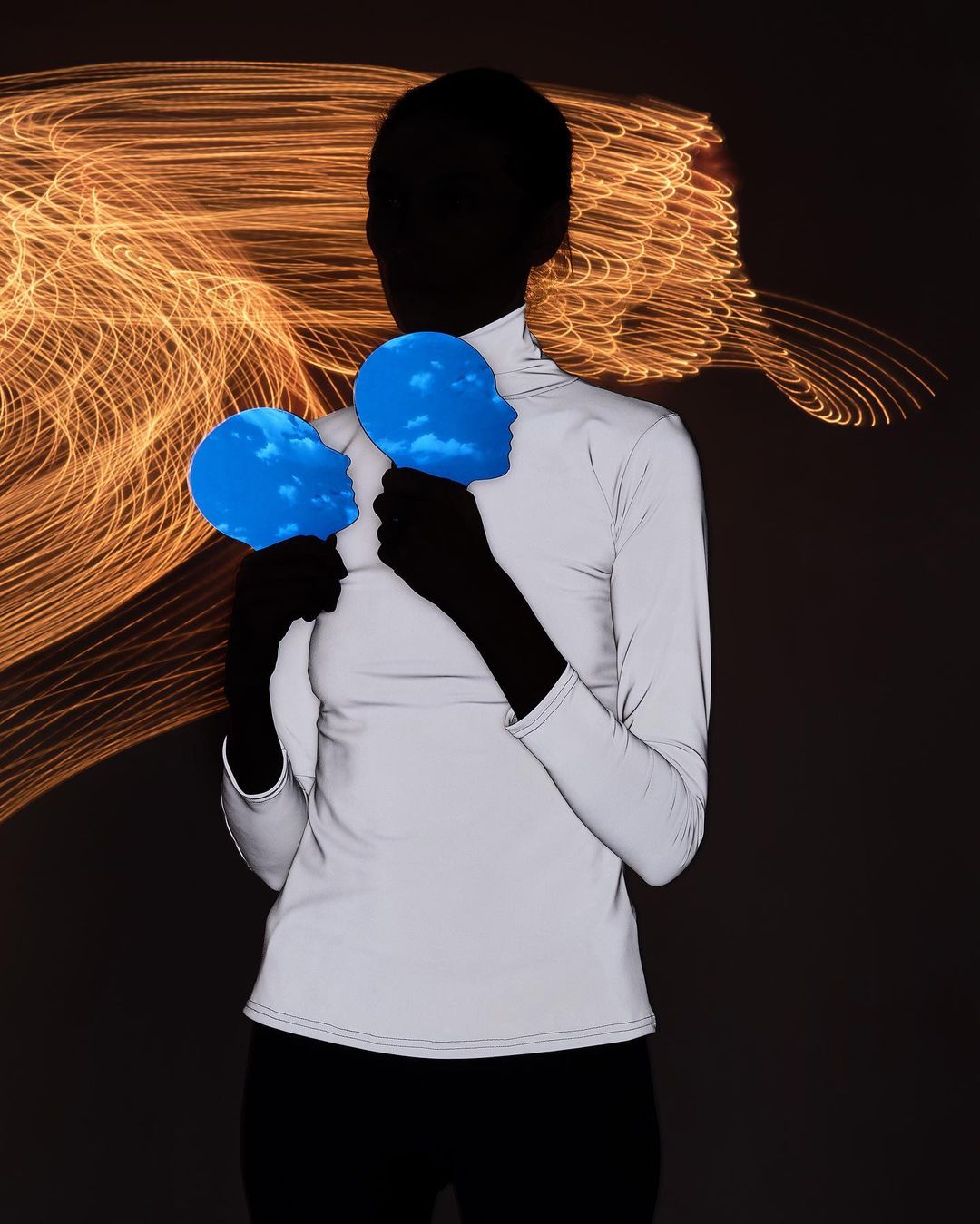 Person holds two face-shaped objects with clouds in front of the body. The background features abstract light trails. Neutral tones prevail.