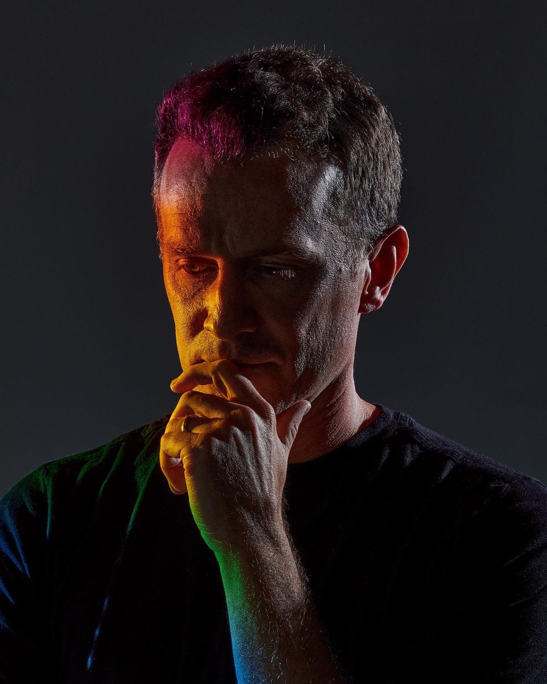 Portrait of a man with hand on chin, illuminated by vibrant multicolored lighting in a dark photo studio setting. 