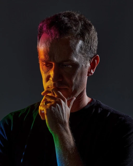 Portrait of a man with hand on chin, illuminated by vibrant multicolored lighting in a dark photo studio setting. 