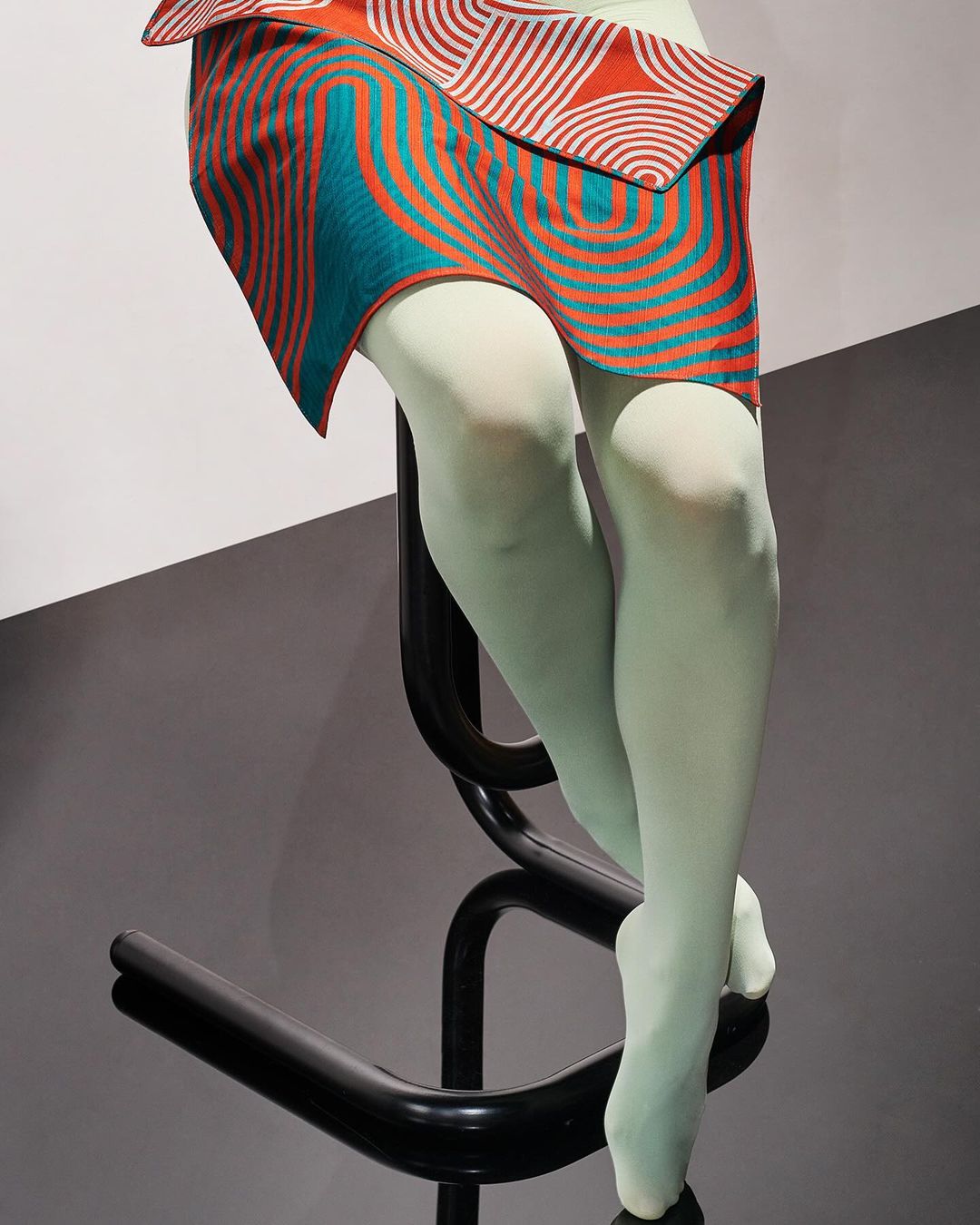 Abstract fashion photo of a mannequin wearing a patterned mini skirt sitting on a black metal chair with a white and gray background.