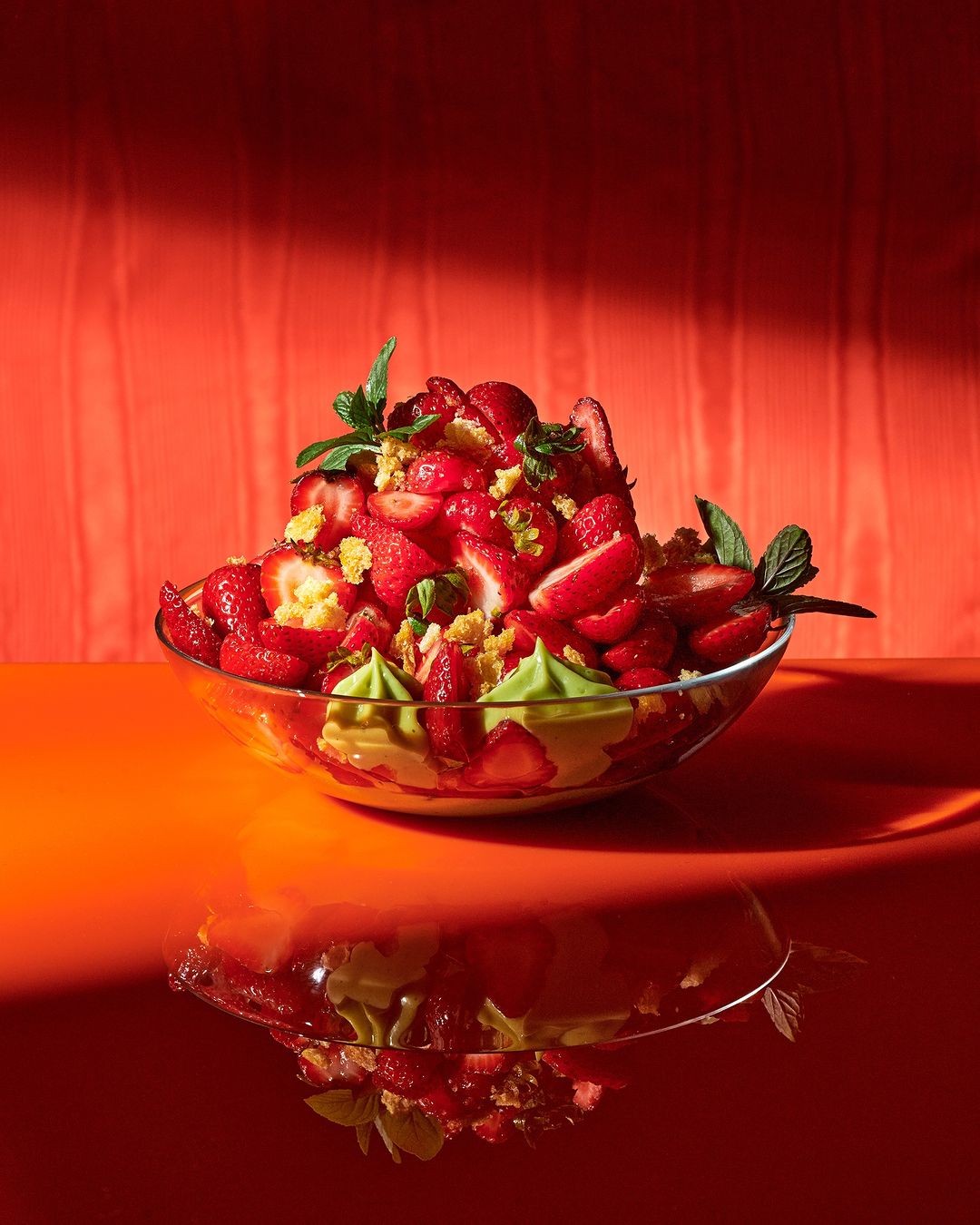 Bright food still life of strawberry dessert in a glass bowl against an orange background with a reflection of the bowl on the surface.