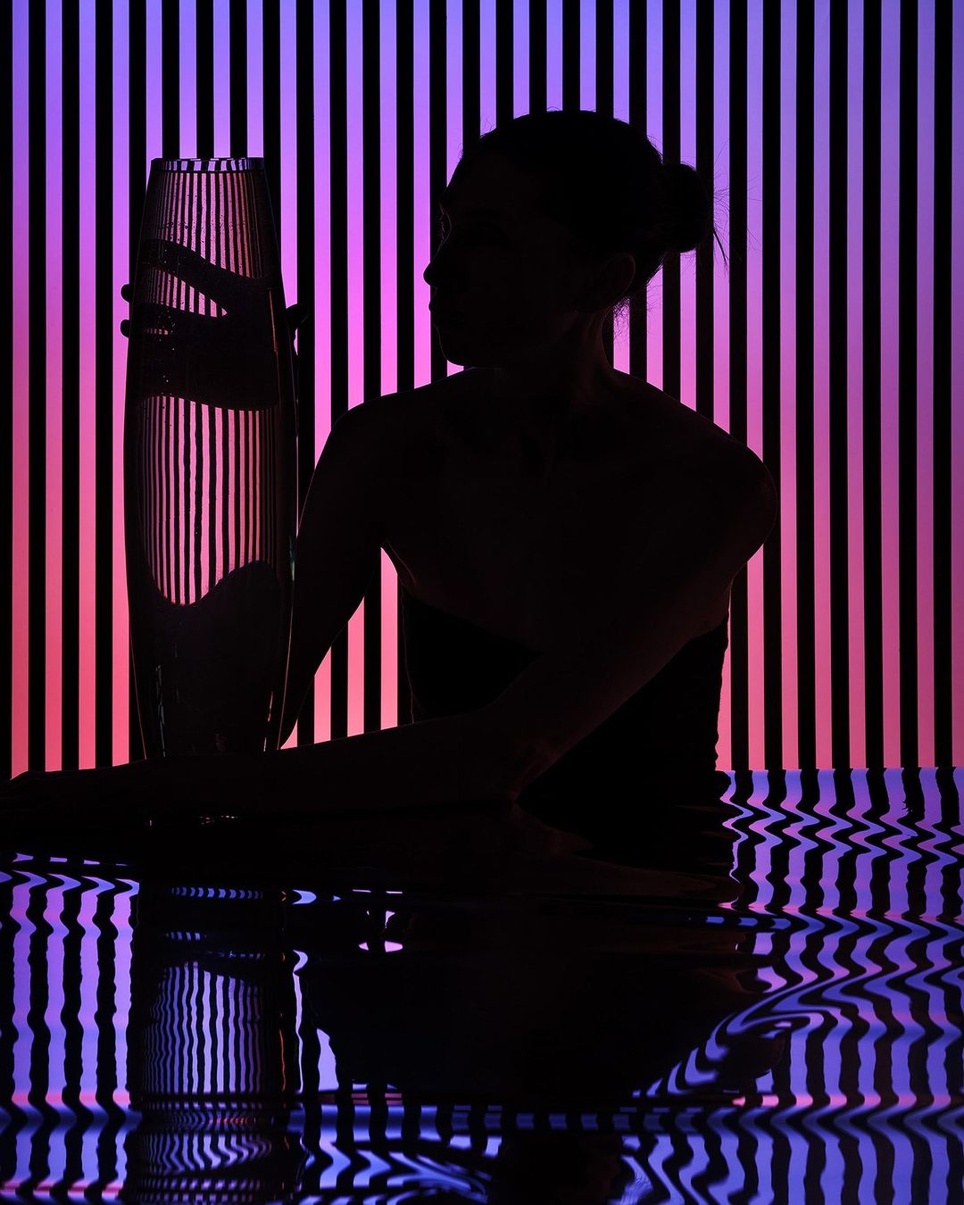 Silhouette of an adult woman holding a glass vase against a striped purple and pink backdrop, creating a stark contrast of light and shadow.