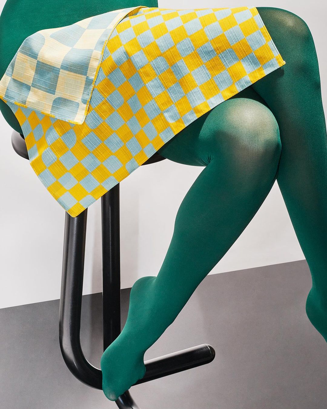 Person wearing green tights sits on a black stool. A blue and yellow checkered cloth is draped over their lap. Minimalist studio setting.