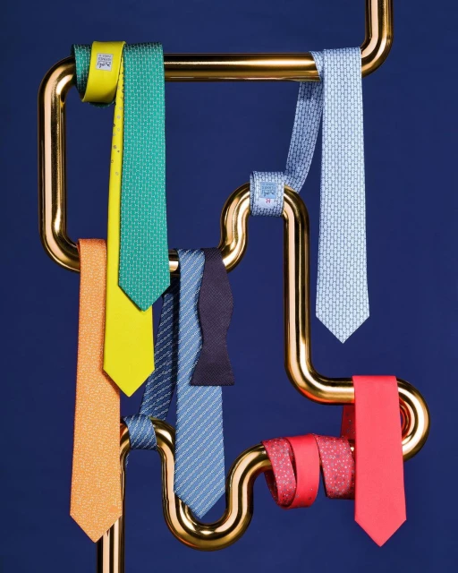 Colorful silk neckties and a bow tie are displayed on a gold-tone metal rack against a dark blue background in a studio setting.