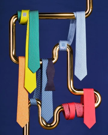 Colorful silk neckties and a bow tie are displayed on a gold-tone metal rack against a dark blue background in a studio setting.