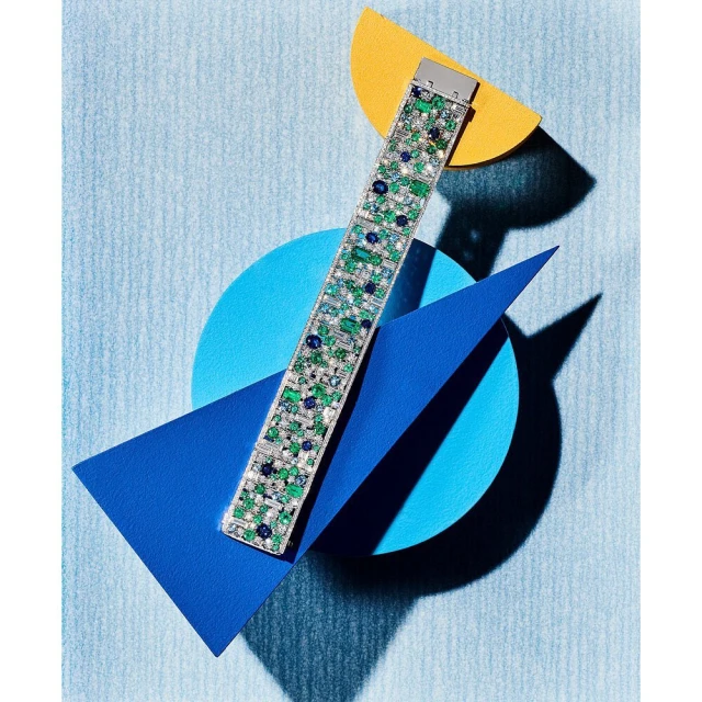 Emerald and sapphire tennis bracelet against a geometric paper background in yellow, white, and shades of blue, lit with studio light.