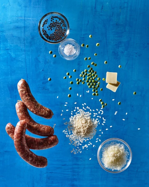 Overhead shot of sausage links, green peas, rice, grains, butter cubes, and shredded cheese ingredients arranged on a blue background.