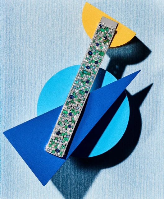 A diamond, emerald, and sapphire bracelet is displayed on a blue surface with geometric shapes in blue and yellow.