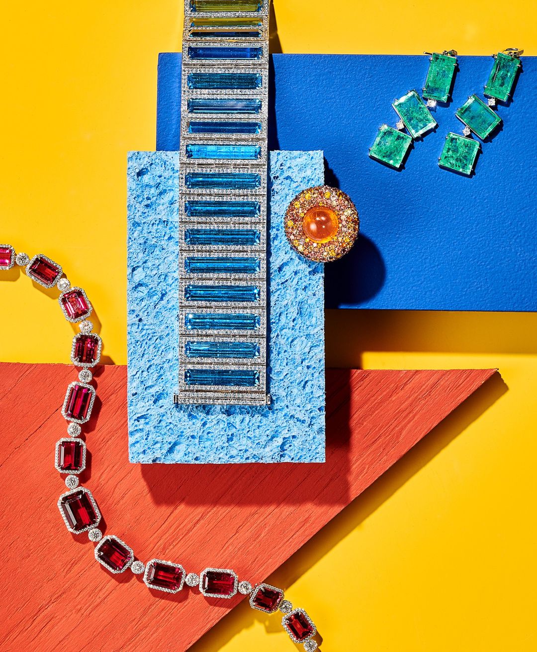 High-angle shot of luxury jewelry displayed on colorful geometric platforms. A necklace, bracelet, ring, and earrings are featured.