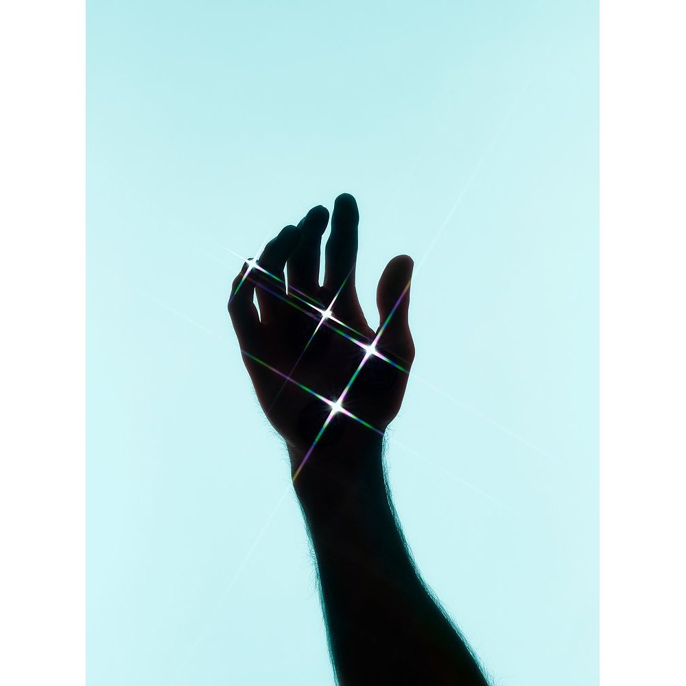 Silhouette of a raised arm and hand with fingers extended against a light blue background, with light flares on the hand.