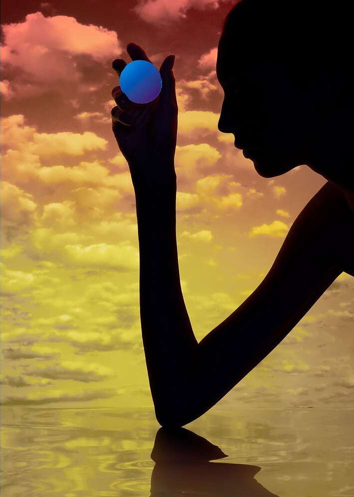 Silhouette of a woman holding a glowing blue orb against a sunset sky with a water reflection, creating a surreal and artistic composition.