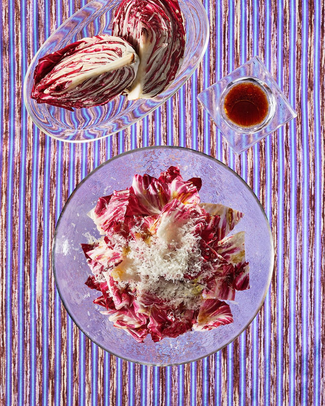 Overhead shot of radicchio salad in a glass bowl with dressing and loose radicchio leaves, presented on a blue striped tablecloth.