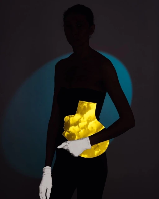 Artful studio shot of a silhouetted adult woman in black holding a yellow, vase-shaped cut-out with cloud imagery, wearing white gloves.