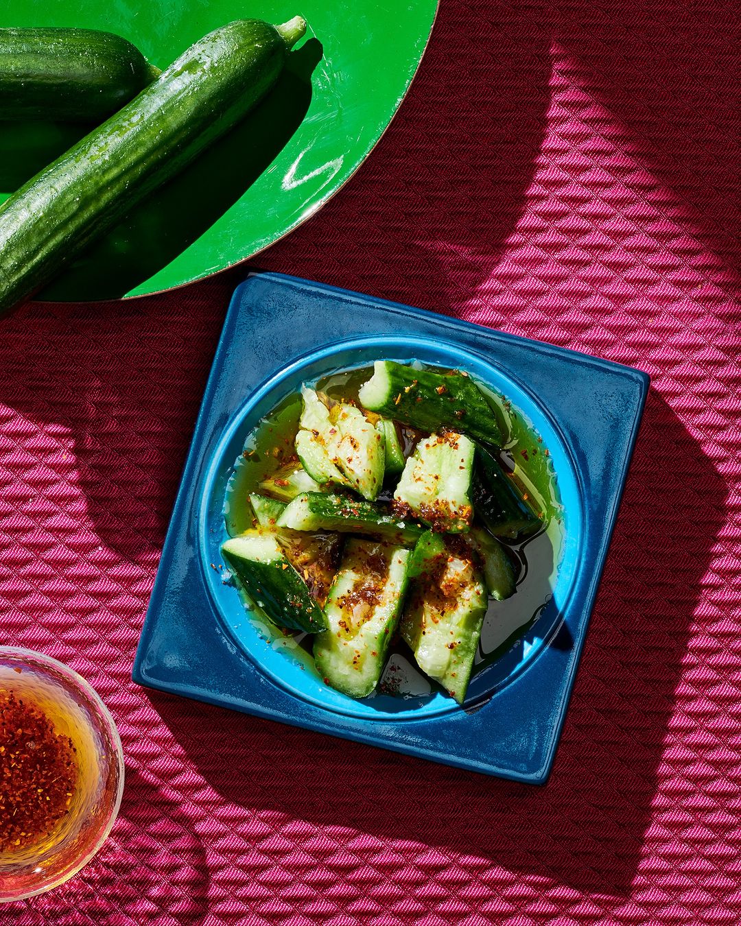 Overhead shot of sliced cucumbers in a blue dish with sauce, arranged with whole cucumbers and a dipping sauce on a pink textured surface.