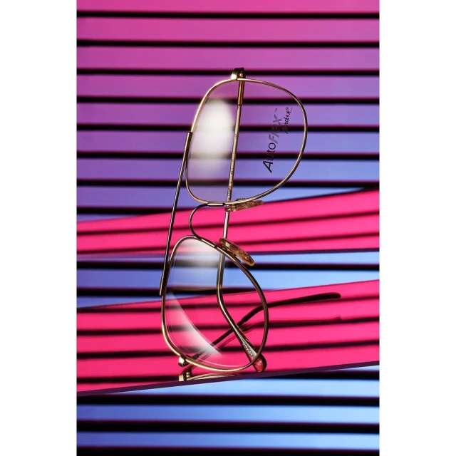 Elegant gold eyeglasses with clear lenses are displayed against a vibrant striped background in pink and blue, creating a stylish eyewear product shot.