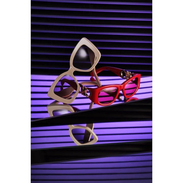 Still life shot of cream-colored, red-framed cat eye sunglasses with purple tinted lenses on reflective surface against purple venetian blinds.