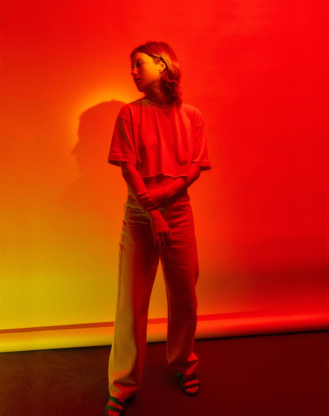 Full-length shot of a woman in a cropped top and jeans, posing against a vibrant red and yellow gradient background, bathed in colorful lighting.