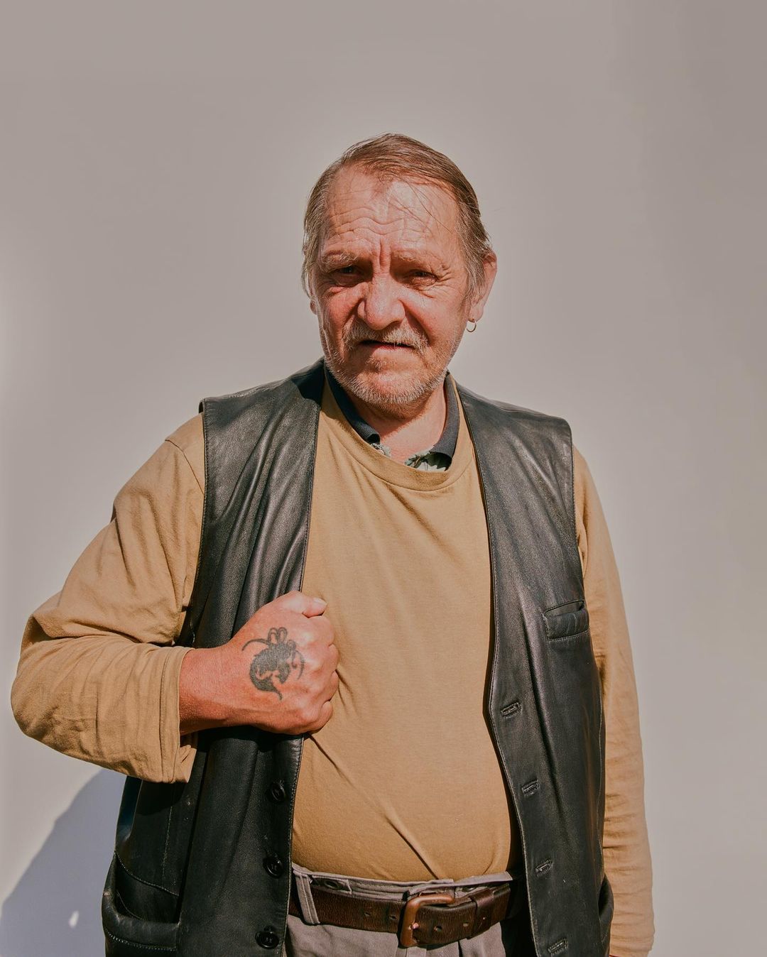 Portrait of an adult male wearing a leather vest and brown shirt with a tattoo on his left hand against a plain white background.