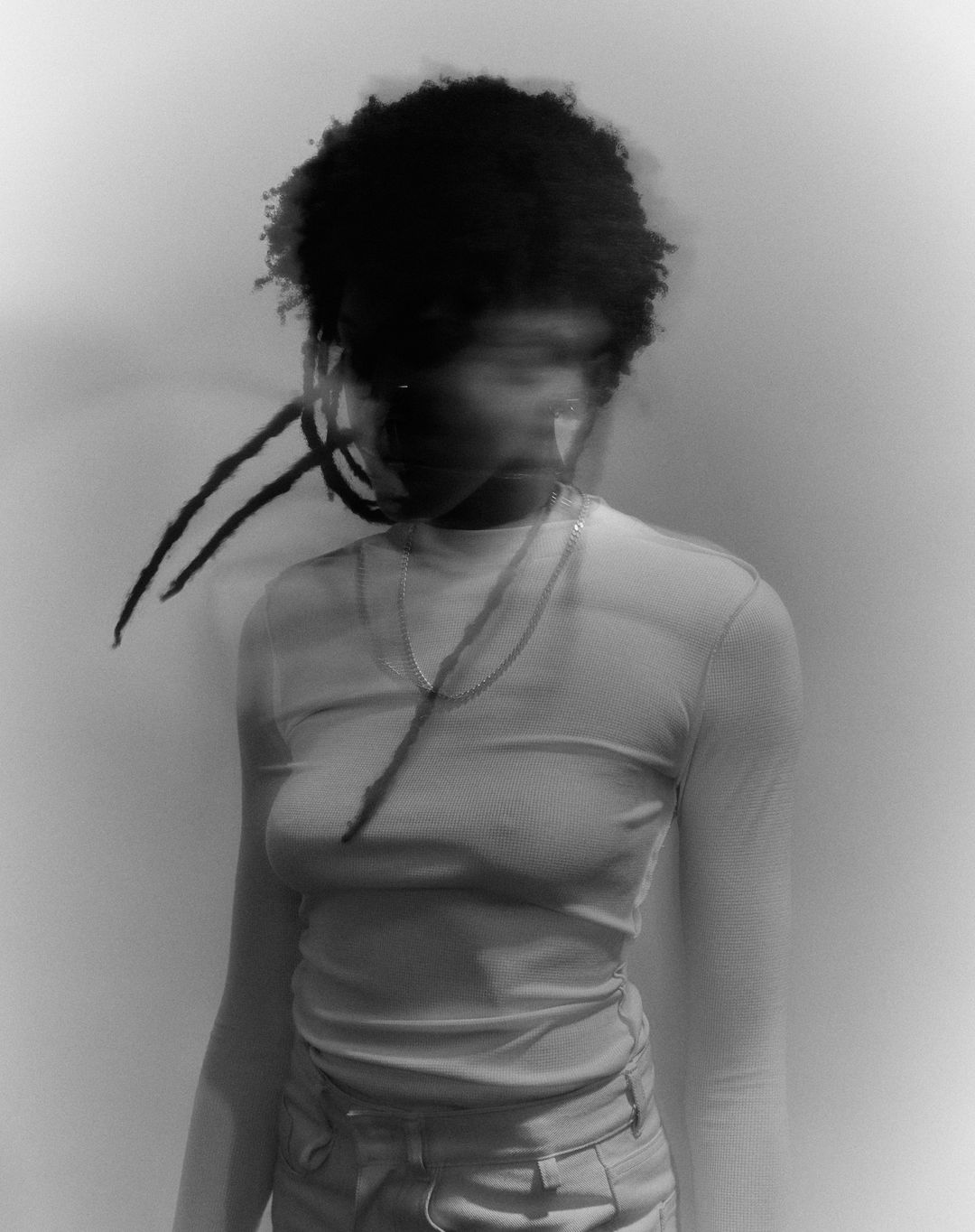 Portrait of a Black female adult in long-sleeved shirt and pants with long dreadlocks posing in a blurred, black and white studio portrait.