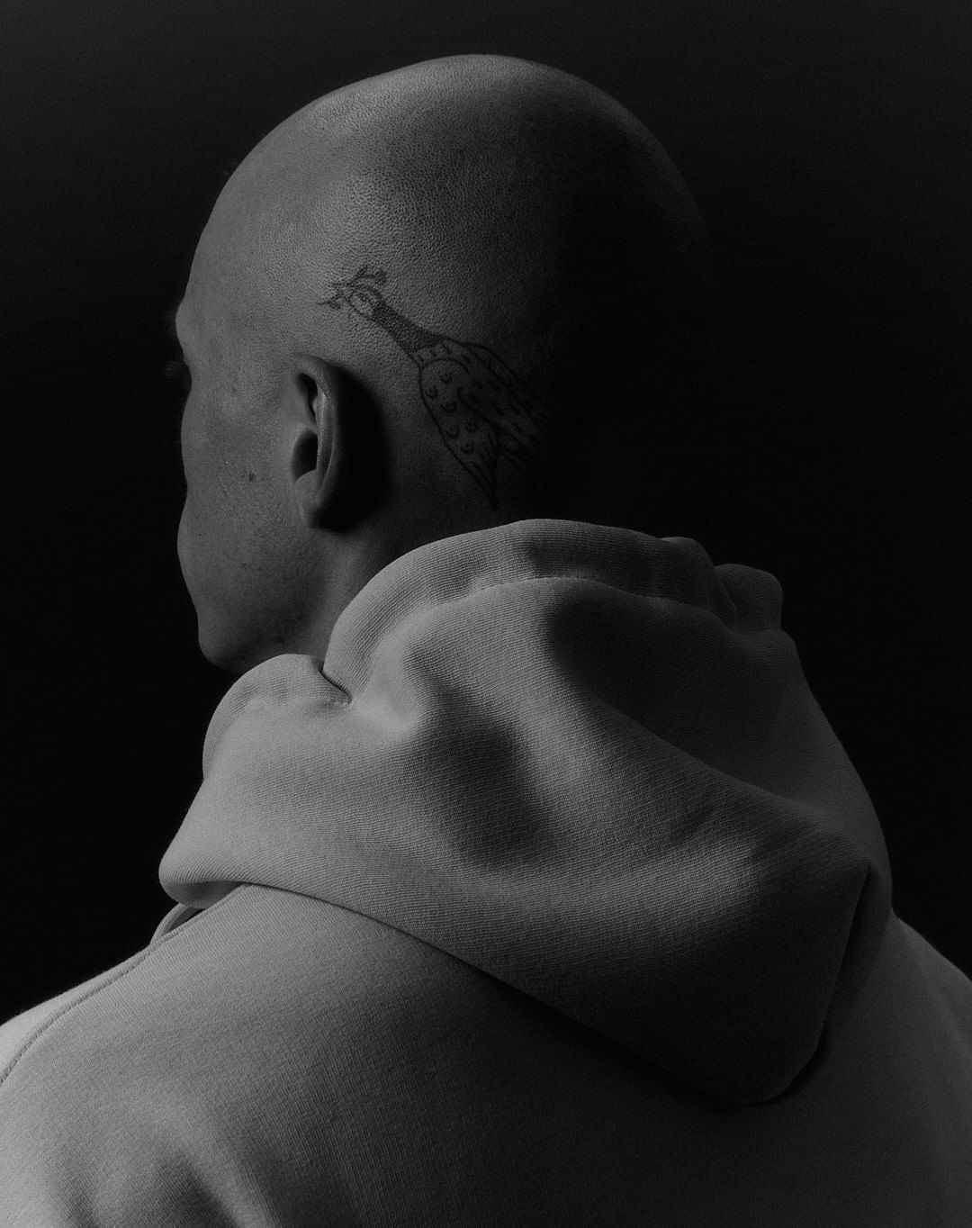 Monochromatic portrait of a bald adult male in a hoodie with a bird tattoo on his head, set against a dark, blurred background.