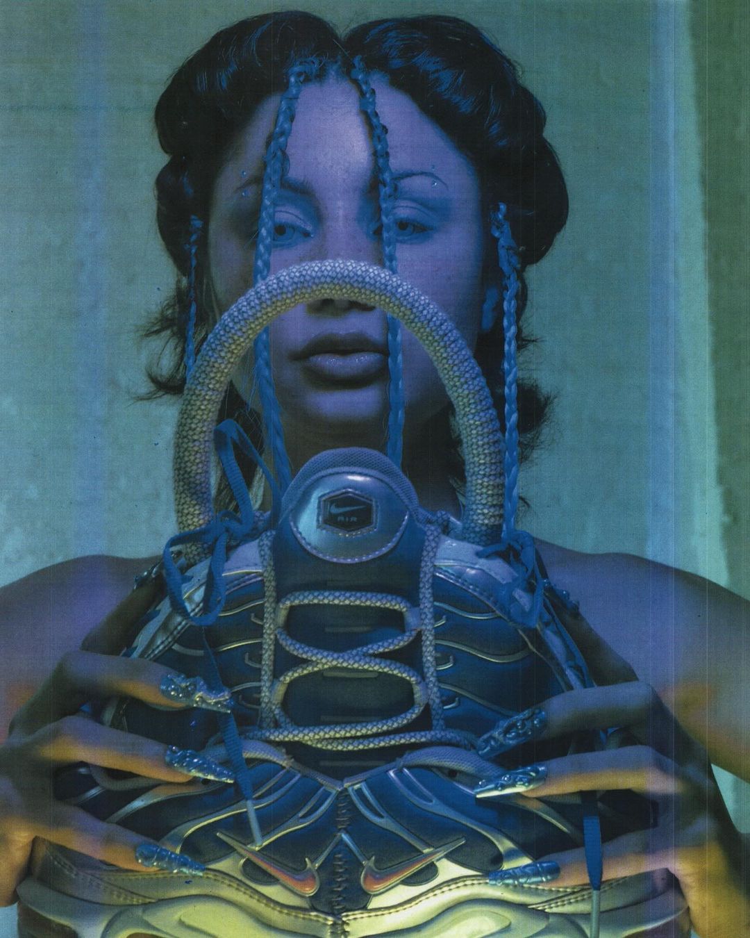 Close-up of a person with long braids posing with a metallic blue and silver Nike Air Max Plus sneaker, indoors, blue-toned.