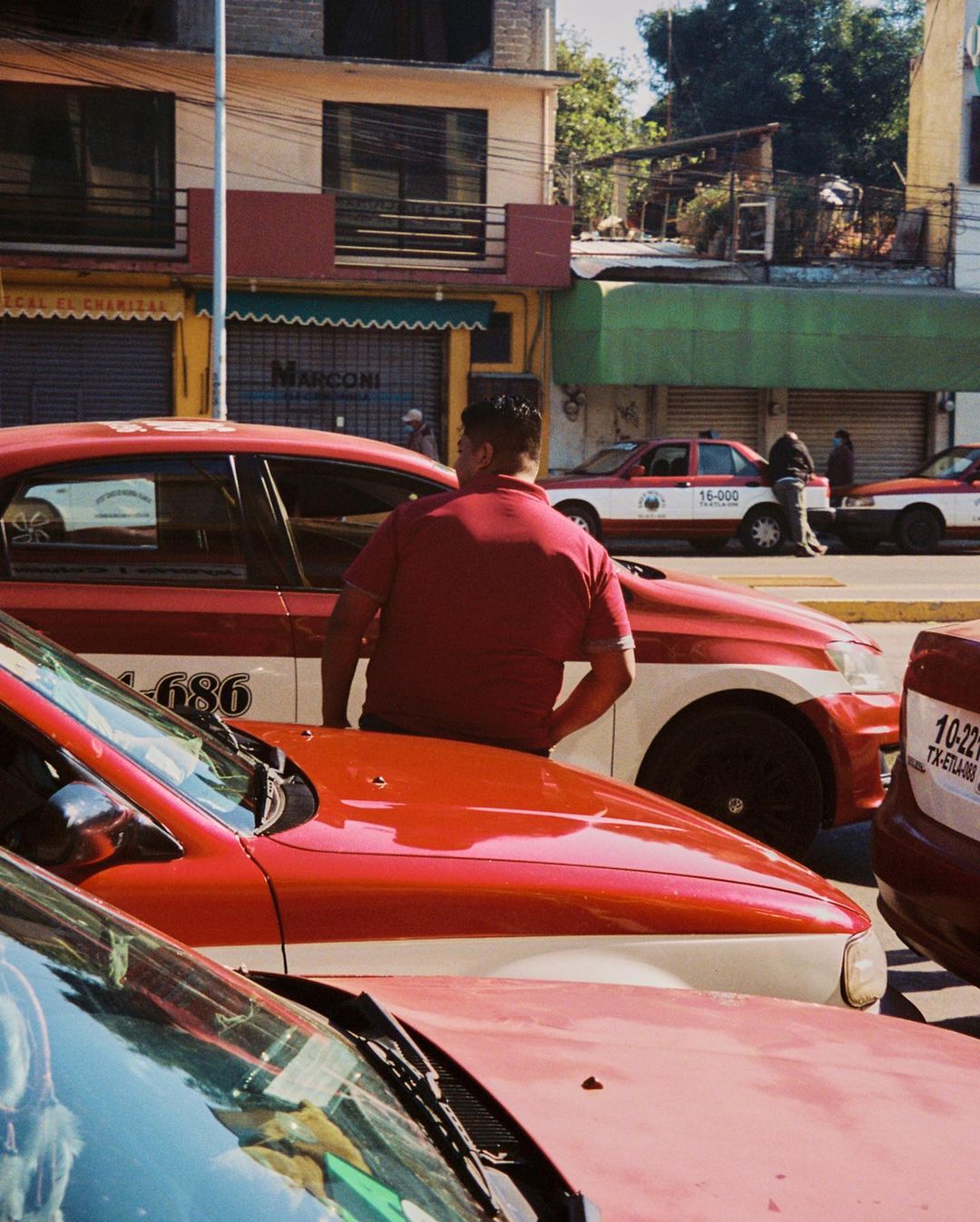 A group of red and white taxis sit on a sunny city street. An adult male wearing a red shirt stands next to one of the taxis.
