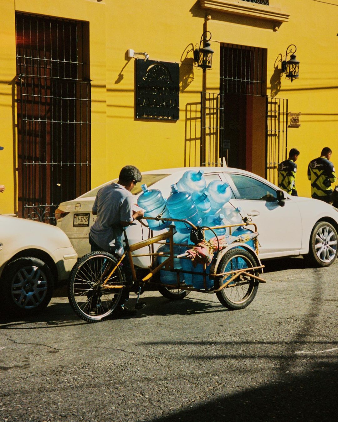 Man transports large water bottles using a three-wheeled bicycle on a city street, with cars and a yellow building in the background.