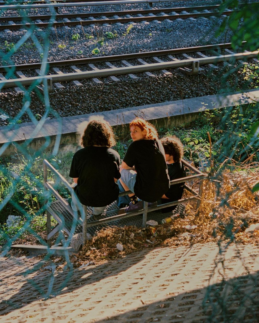 Three people sit on metal stairs near railroad tracks. One person faces forward with red hair, while two people face away from the camera.