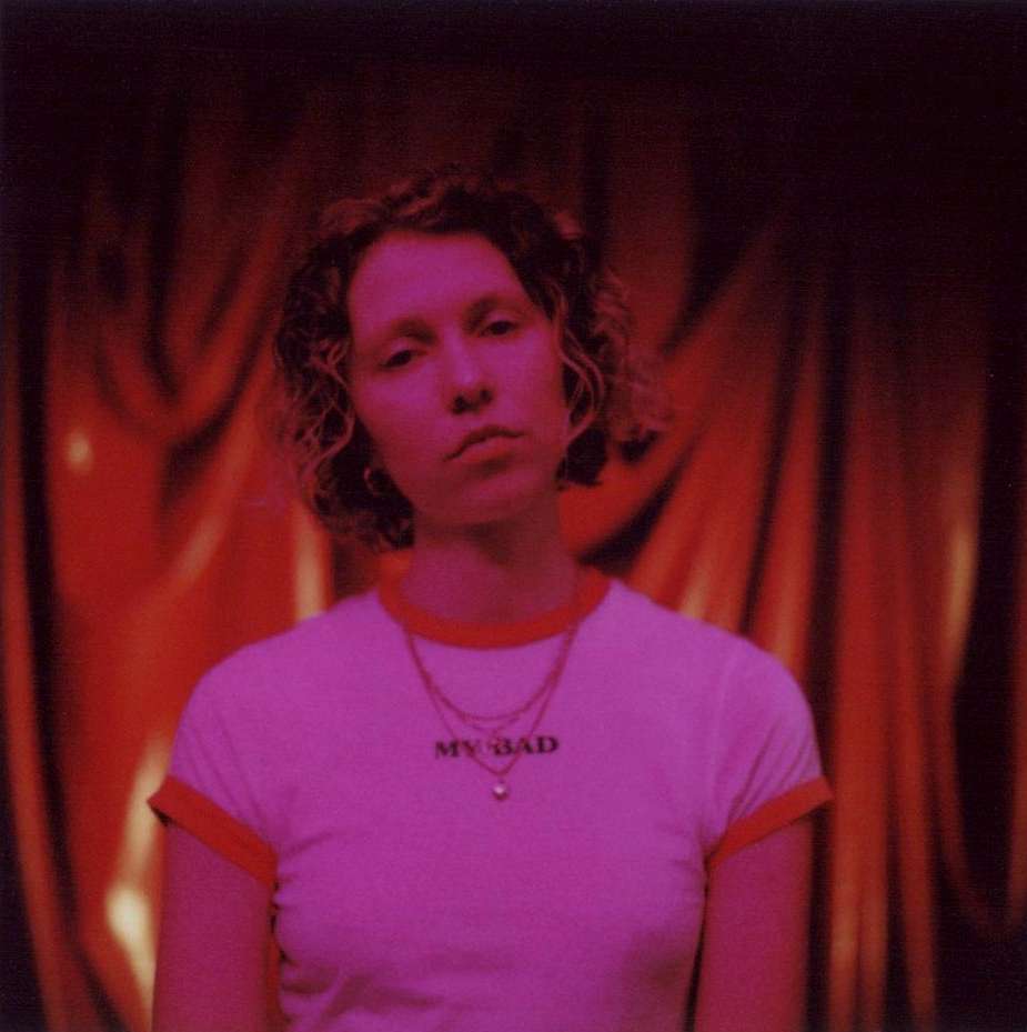 Portrait of a young adult woman with short curly hair wearing a 'My Bad' t-shirt in a dimly lit studio with red curtains.