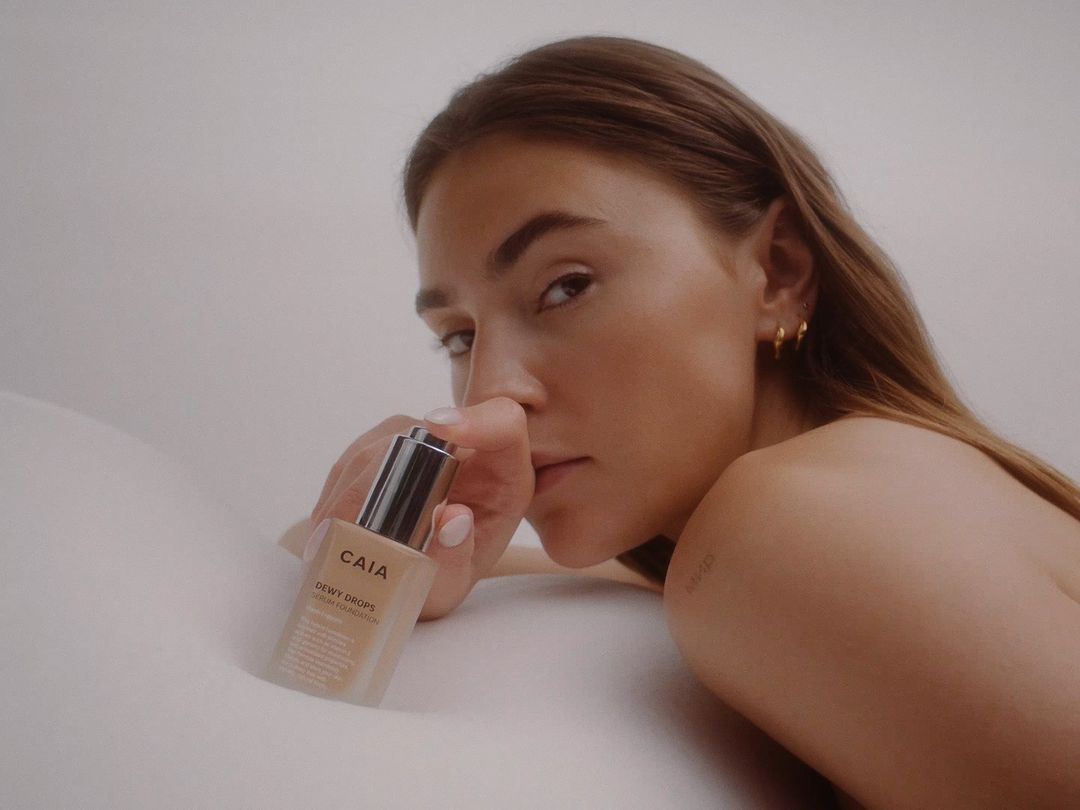 A woman poses with a bottle of CAIA Dewy Drops serum foundation against a light background in a beauty or skincare advertisement.