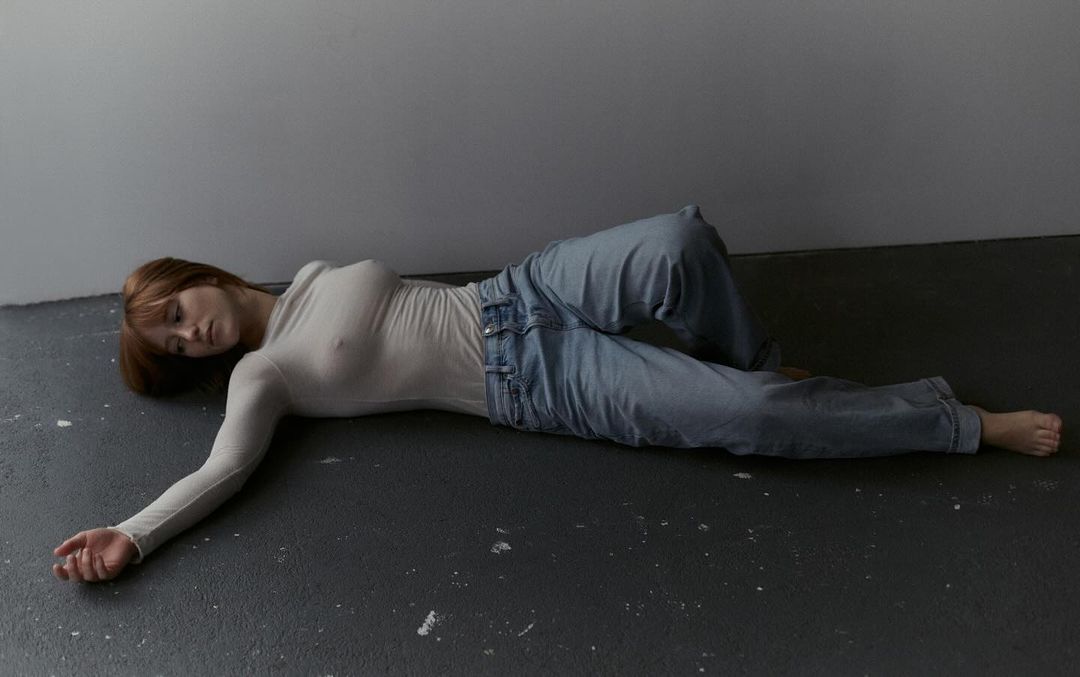 Woman with red hair lying on a grey floor wearing jeans and a long-sleeved white shirt in a minimalist, contemporary setting.