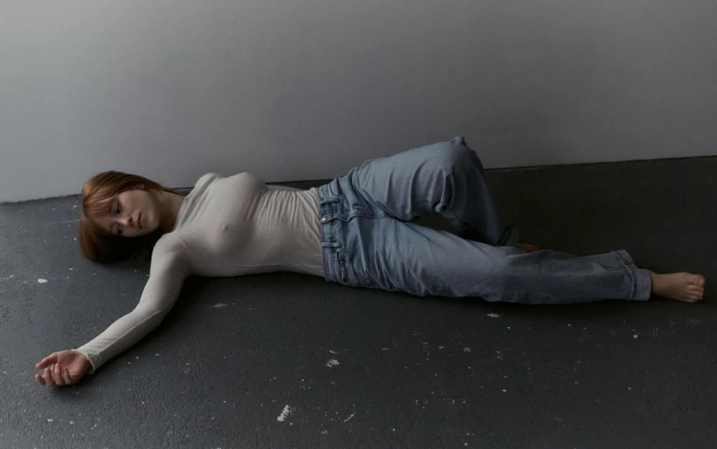 Woman with red hair lying on a grey floor wearing jeans and a long-sleeved white shirt in a minimalist, contemporary setting.