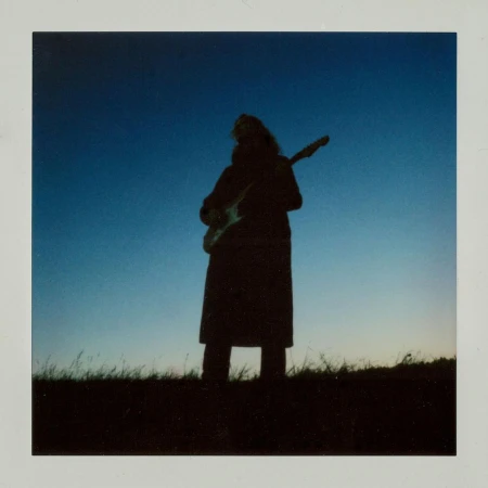 Silhouette of a person with long hair standing outdoors holding an electric guitar against a gradient blue sky and grassy field.