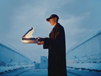 Man wearing a dark trench coat and cap holding a burning box outdoors against a pale blue sky and infrastructure walls