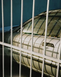 Close up view of a dusty white car parked behind vertical white metal bars casting dramatic shadows