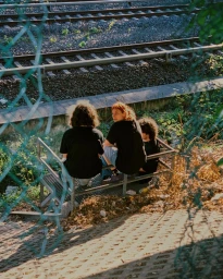 Three youths sitting together on a metal staircase outdoors near railway tracks, viewed through a green chain-link fence.