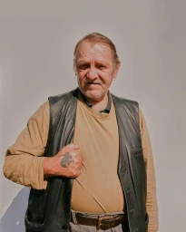 Portrait of an older man with a hand tattoo wearing a leather vest and beige sweater against a neutral background.