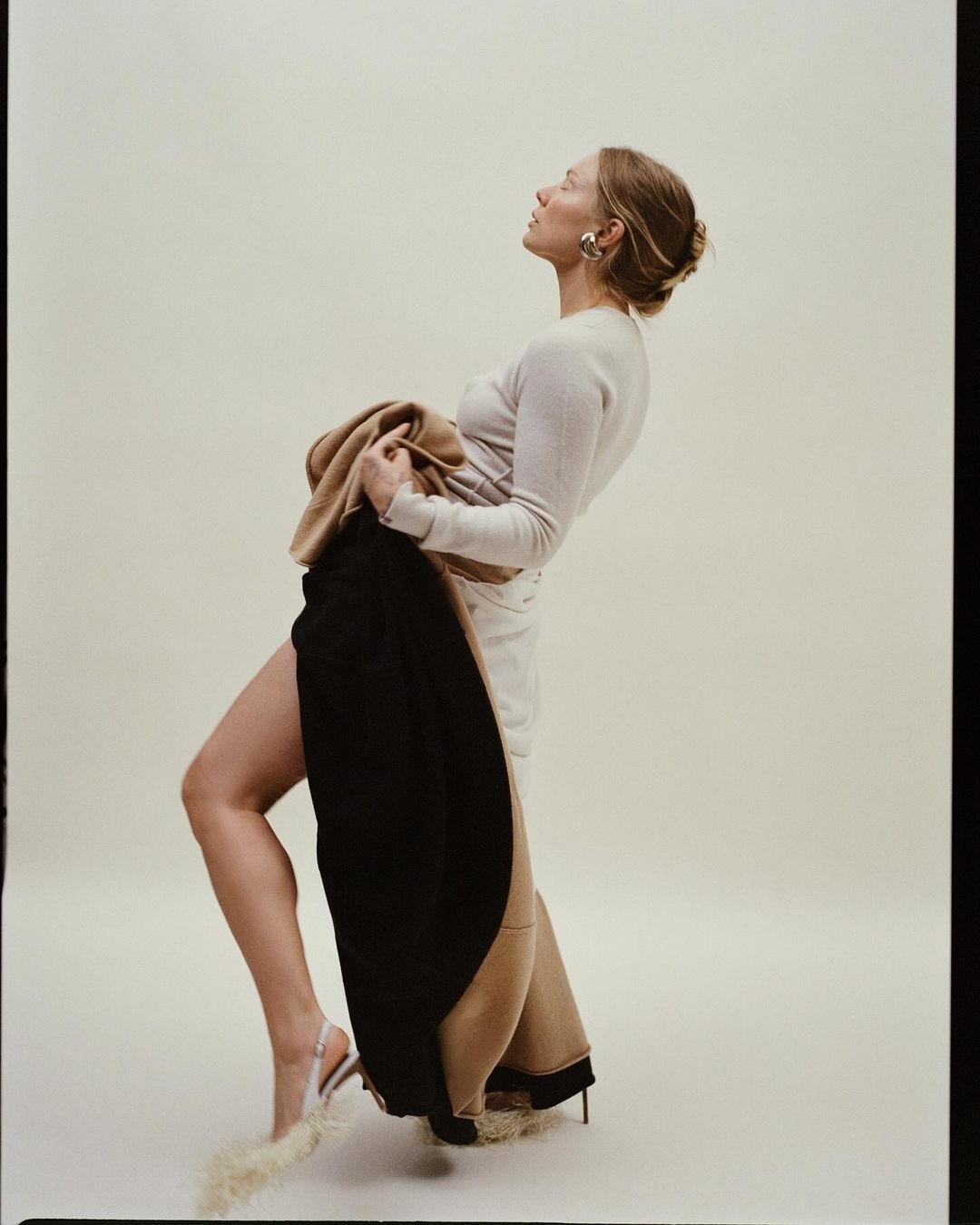 Woman poses in a light-beige long-sleeve dress, holding a black and beige coat, with fur-embellished high heels, in a photo studio.