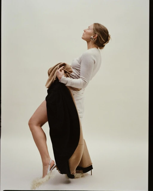 Woman poses in a light-beige long-sleeve dress, holding a black and beige coat, with fur-embellished high heels, in a photo studio.