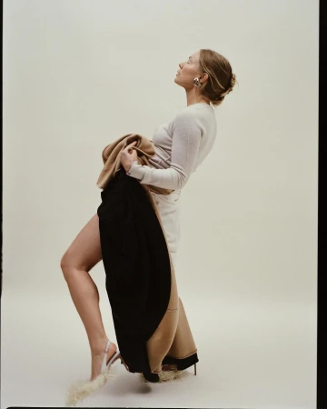 Woman poses in a light-beige long-sleeve dress, holding a black and beige coat, with fur-embellished high heels, in a photo studio.