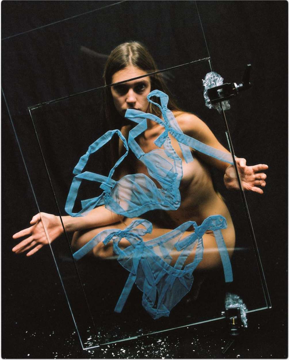 A striking studio portrait captures a nude adult woman crouching behind a clear glass pane with blue tulle fabric in a stark black environment.