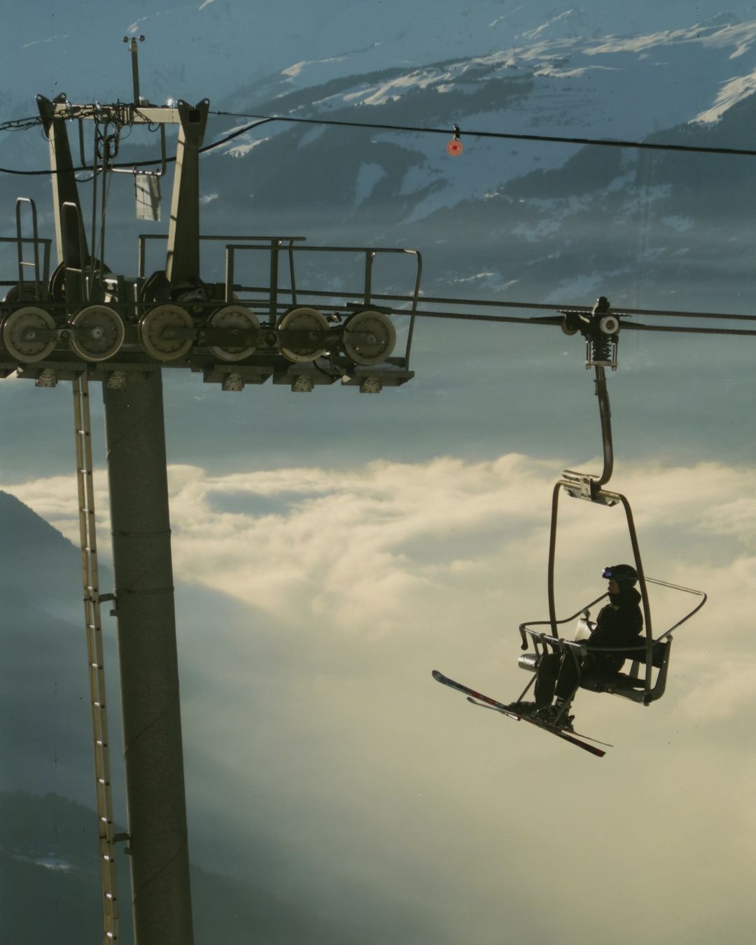 Skier rides a ski lift up a mountain on a sunny day with cloud cover, with ski lift mechanism visible.