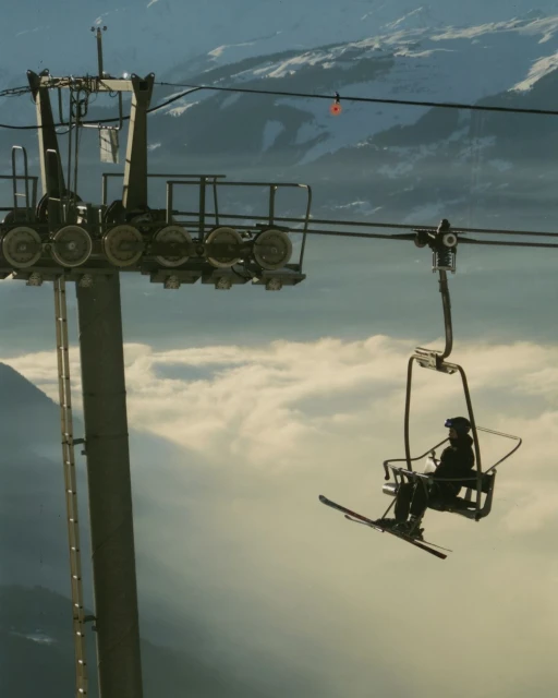 Skier rides a ski lift up a mountain on a sunny day with cloud cover, with ski lift mechanism visible.