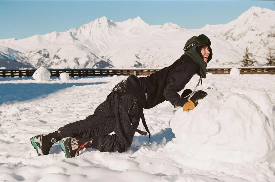 Adult female in snow gear pushing a large snowball in a snowy mountain environment under a clear blue sky on a sunny day.
