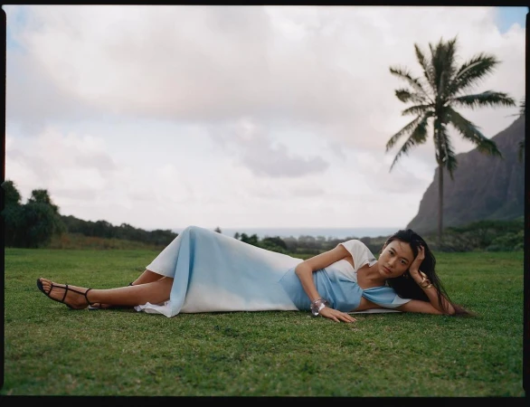 Model lying on grass wearing a blue and white dress with a palm tree and ocean in background