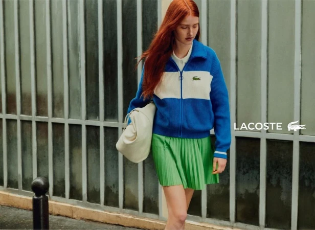 Red-haired female model wearing blue and white zip cardigan and green pleated skirt holding a cream handbag outside a building.
