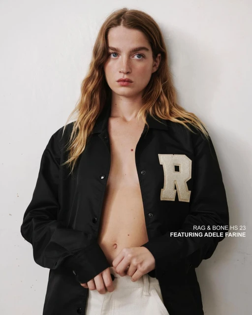 A young woman with wavy blonde hair poses in a black jacket and off-white pants against a white wall in a studio fashion shot.