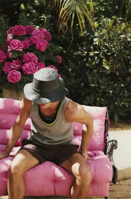 Man in tank top and shorts sits on pink cushioned bench in a garden setting with green foliage and pink flowers in the background.