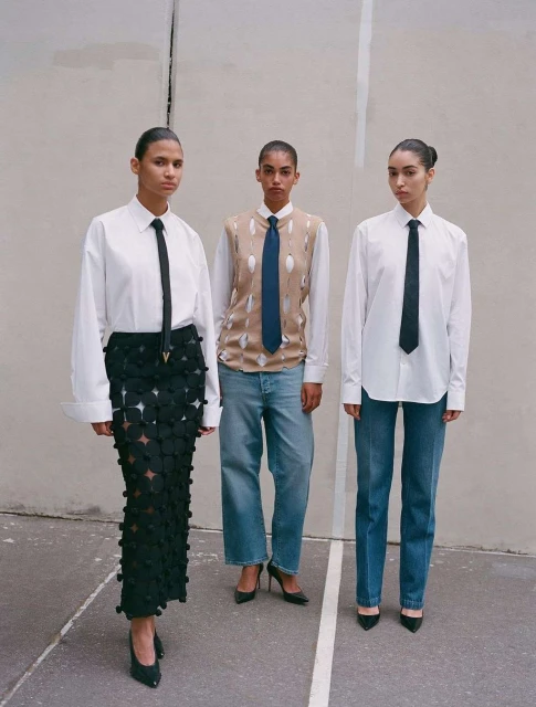 Three women in fashionable business attire, wearing button-down shirts, ties, and unique skirts and jeans, posing indoors against a wall.