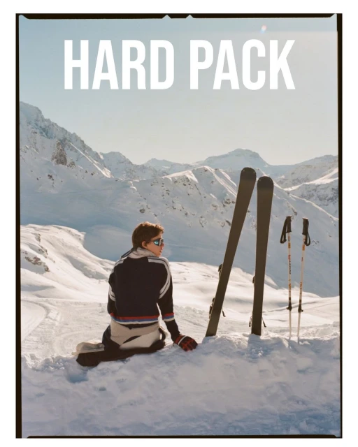 Person wearing ski apparel sitting in snow with skis and poles; snowy mountain landscape against a bright, sunny sky in the background.